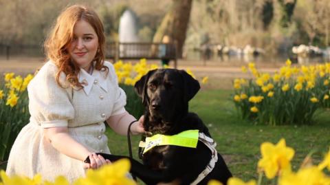 Emily, a young woman with mid length auburn hair, kneels in a park beside Rosie, a black Labrador guide dog, gently holding her paw. Emily is wearing a light cream dress and sitting among bright yellow daffodils, with flowers in front of her and behind her. Behind them is a large tree trunk, a fountain, and out of focus greenery.