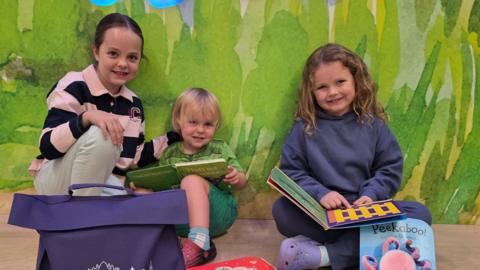 Three children of different ages are sat on the floor, some holding books. They are in a colourful room with a wall that has some artwork on it behind them. They are all smiling at the camera.