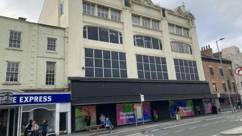 The former Debenhams department store in Stockton. It is a multi-storey building, painted cream with the majority of its windows blacked out. The lower area is painted black and is adorned with signs reading "Welcome to Stockton". Someone is sitting on a bench on the pavement, while three people stand chatting on the street nearby.