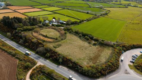 An ariel shot of Castilly Henge which is at the centre of a large field. The henge is a circle. To the left of the large site, there is a road. On the right of the henge, there are many green and yellow fields. 