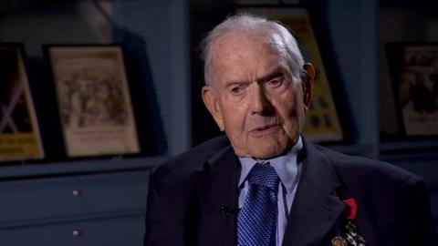 Percy Chafer, an elderly man in a light blue shirt and tie and navy blue jacket featuring military medals. He sits in a blue room at the National Archives with framed historical posters behind him.