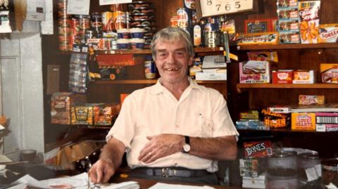 Dave Masters with grey hair and a pale cream shirt is sitting at the counter in his village shop, smiling at the camera. There are sweets and other food items stacked on shelves behind him. The photo was taken a few decades ago, with old branding visible on Quality Street boxes, Fruit Pastel packets and Fray Bentos pies, among other items.