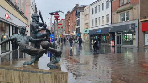 Statue of cricketer, footballer and rugby player on plinth with wide wet pavement and shops on both side with red lanterns across the street 