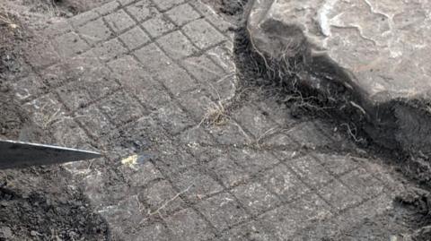 The Roman board game when it was still in the ground. It is a grey slab of rock with a grid pattern. Someone is holding a knife to the left of the photo and a large grey rock to the right.