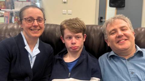 A woman, a man and a young man sit close together on a brown leather sofa.