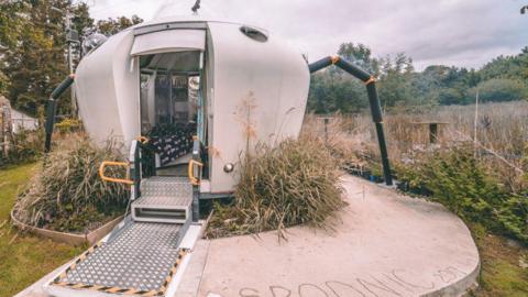 A white UFO-like building sat amongst overgrown plants. On the tarmac in front, writing engraved reading 'Spodnic 2017'. The door opens out front with a metal walkway and two steps leading inside the pod. A grey cloudy day lies behind.
