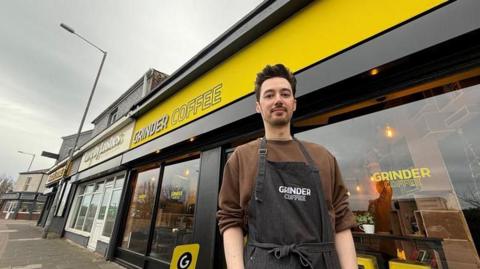 Riki Tsang has gelled up black hair and stubble, and wears a black apron saying "Grinder Coffee" outside yellow cafe with the sign "Grinder Coffee".