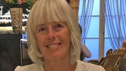 Ann Blackwood is pictured smiling in a room with French windows behind her. She has short white hair with a fringe and wears a white top.
