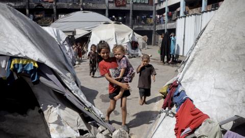 A smiling older child carries a younger girl in her arms in a displaced persons camp in Khan Younis, Gaza. The pair are surrounded by dirty grey and white tents with items of clothing drying on the outside. there are other children and women in background.