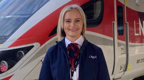 A blonde woman in a uniform standing on a train station platform in front of an LNER Azuma train. The train features a red, yellow, grey, and white colour scheme and is positioned along the edge of the platform under overhead power lines. The person is wearing a dark blue uniform jacket with a visible LNER logo on the chest, along with a red necktie or scarf. They are holding a framed certificate which includes a large red star.