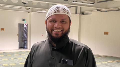 Abdul Basith is a Muslim man wearing a brown tunic and a white head covering with a beard. He is standing in a large prayer hall and smiling. He has a phone in his pocket