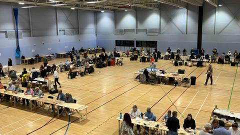 People sitting at tables in blue sports hall counting ballot papers