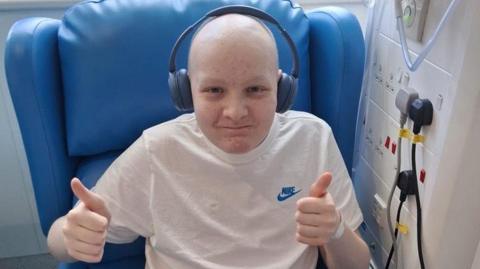 A boy in a hospital chair. He is bald, wearing noise-cancelling headphones, and gives two thumbs-ups to the camera.