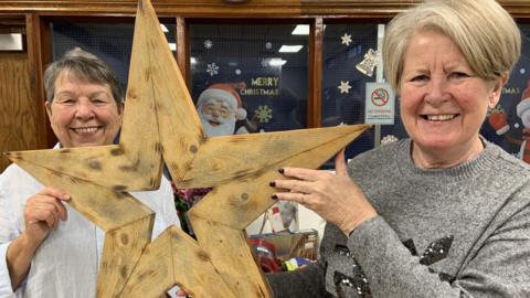 A woman with short dark hair is wearing a white shirt and is stood next to a woman with short blond hair in a grey sequin jumper. They are holding up a large wooden Christmas star