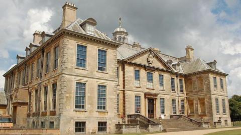 A large historic stone mansion with tall sash windows, ornate architectural detailing and multiple chimneys, set against a dramatic cloudy sky. The building features a central entrance with steps leading up to it, flanked by symmetrical wings. A gravel path curves across the neatly kept lawn in the foreground.