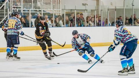 Five ice-hockey players on the ice holding hockey sticks and chasing a puck, with a crowd looking through perspex glass in the background
