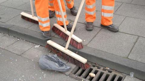 A stock image of three refuse workers in hi-vis trousers using brooms to sweep litter in the street. 