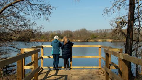Two women stand on the new wooden boardwalk overlooking the lakes. It is a sunny day.
