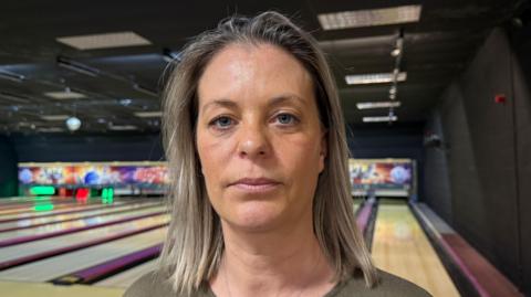 Fiona Watts has light medium length blonde hair and blue eyes. She is in front of the bowling lanes at her bowling alley. In the background red and green lights can be seen illuminating the bowling pins.