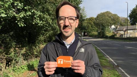 A man in a black coat with glasses holds an orange bus pass.