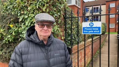 Steve Bryant, an elderly man in a flat cap and tinted glasses, standing in front of a black iron fence and ivy-covered hedge. Behind the fence is a modern three-storey block of red-brick flats.