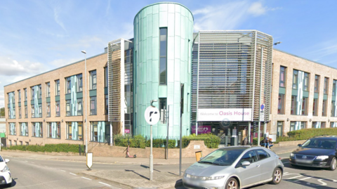 A photo of the front of an L-shaped modern building on a street corner, taken from the middle of the road opposite. A white sign above the doors says Welcome to Oasis House and a pale green cylinder structure forms the corner of the L.  The pale brick building has multiple windows over four floors 