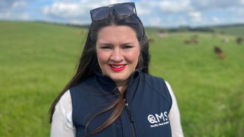 Sarah Millar, wearing a white shirt and blue gilet marked with the QMS logo, looks to the camera at the side of a field.  She has dark brown hair, red lipstick and has sunglasses on her head.
