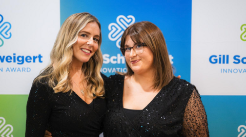 Leah and Florence are both wearing black, shimmery dresses and are stood in front of a blue, white and green advertisement board. Leah has long, wavy blonde hair and Florence has bobbed brown hair and glasses.
