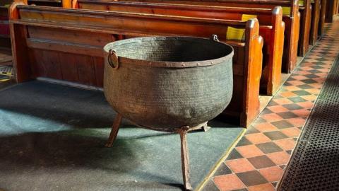 A large black copper cauldron on a rusty metal stand with three legs. It is in the nave of a church. In the background are rows of pews