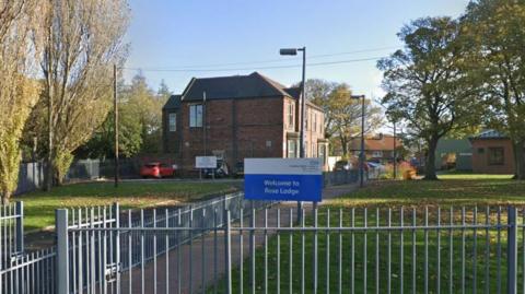 Rose Lodge in Hebburn. The ward is a converted detached house. A paved path leads to the front doors of the property. Green lawn lines either side of the path. The building is located behind a metal fence and a welcome sign.