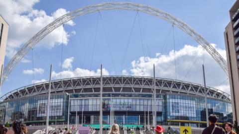 People gather outside Wembley Stadium, with its arch, on a sunny day.