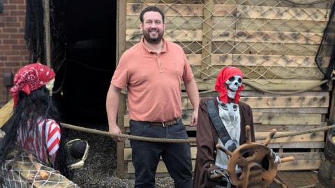 A man stands outside next to two models of skeletons. The man is smiling and he has brown hair and a brown beard. He is wearing an orange t-shirt and grey trousers. The skeleton models are dressed as pirates, with red bandannas, and one has been put next to a ship's steering wheel.