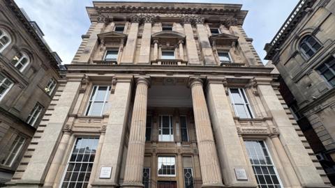 The Old Post Office building. It has a Victorian structure, is made from sandstone, and has a classical style. There are two pillars that line the entryway, and there are tall glass windows to the other side of each pillar. It is a multi-storey building.