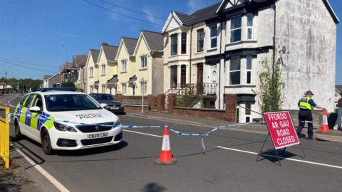 A police car is seen along a road, with police tape and a red "Road Closed" sign