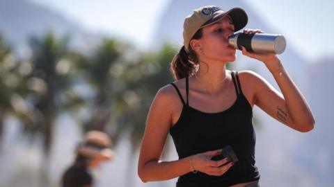 A woman drinks water on Copacabana Beach in Rio de Janeiro, Brazil, during a heatwave. She is wearing a black vest top and a light brown cap. Palm trees are visible in the background.