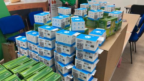 A wooden table stacked high with blue and green boxes containing smart plugs and energy saving lightbulbs.