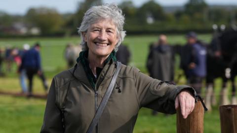 Catherine Connolly wears a green jacket. She has short grey hair and is smiling. She is standing in a field and leaning her arm on a post. 