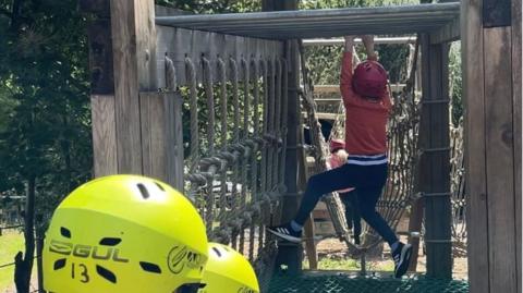 A child wearing an orange jumper and red helmet hangs from some monkey bars. Two children with yellow helmets look on.