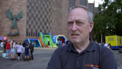 A man with grey hair is standing in front of a large brown brick cathedral and is wearing a dark navy t-shirt with orange writing that reads "feed the hungry"