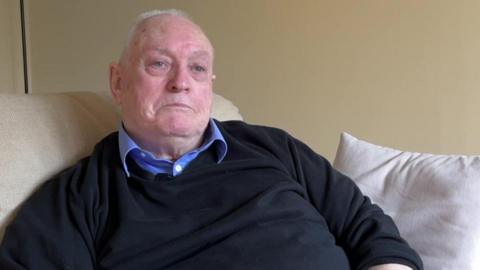 A man sat on a sofa wearing a black jumper and blue collared shirt.