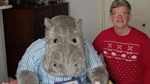 Philip Angier is a man in his 70s wearing a red Christmas jumper with brownish hair and glasses. He is sitting next to a huge hippo soft toy which is almost as big as he is 