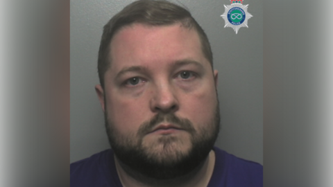 A police mugshot of a man with short dark hair, a beard and a blue shirt.