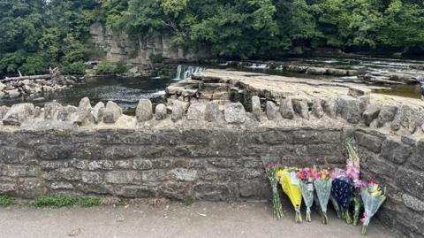 Bouquets of flowers next to a stone wall, with a river flowing in the background.
