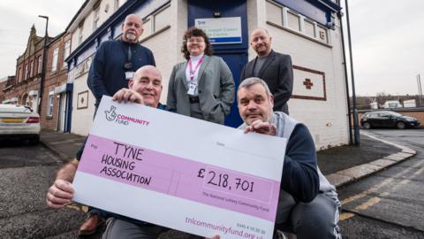 Tyne Housing residents Karl and Rob holding a large cheque from The National Lottery for £218,701. Behind them stands Bryan Beverley from Tyne Housing, Kayah Worrall from The National Lottery Community Fund, and Tyne Housing CEO Steve McKinlay. They are standing outside the Joseph Cowen Centre in Byker.