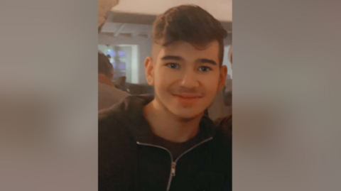 A man wearing a black zip up hoodie smiles into the camera.