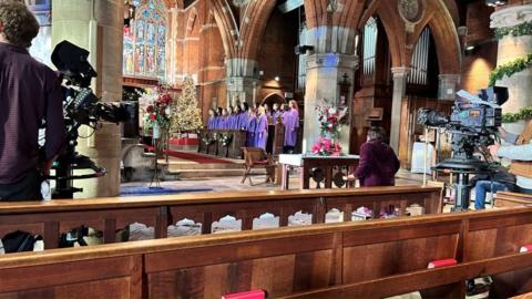 A church with a man and a camera in the foreground with a choir in purple robes in pews in the background. The church has ornate arches and stained glass windows.