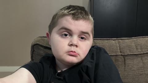 Harry, 11, with short dark blonde hair, sits on a sofa. He is wearing a black t-shirt.