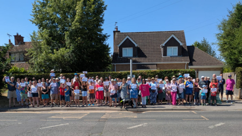 A group of people standing on a roadside in Caversham protesting the bus route closure on a fine summer's day.