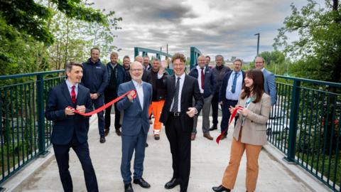 Four smartly dressed people hold red ribbons and smile while stood on a footbridge. A larger group of people, almost all of them men, standing behind them in the background.