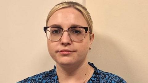 Sophie has blonde hair and wears glasses. She is wearing a blue blouse and looking at the camera. 